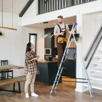 Artisan du bâtiment sur un escabeau expliquant le projet pour rénover sa maison à Montpellier à une cliente dans un salon moderne