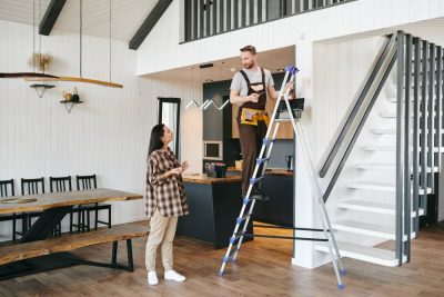 Artisan du bâtiment sur un escabeau expliquant le projet pour rénover sa maison à Montpellier à une cliente dans un salon moderne